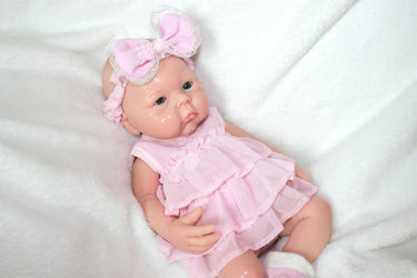 A baby doll dressed in a pink dress and matching headband, sitting against a soft background.